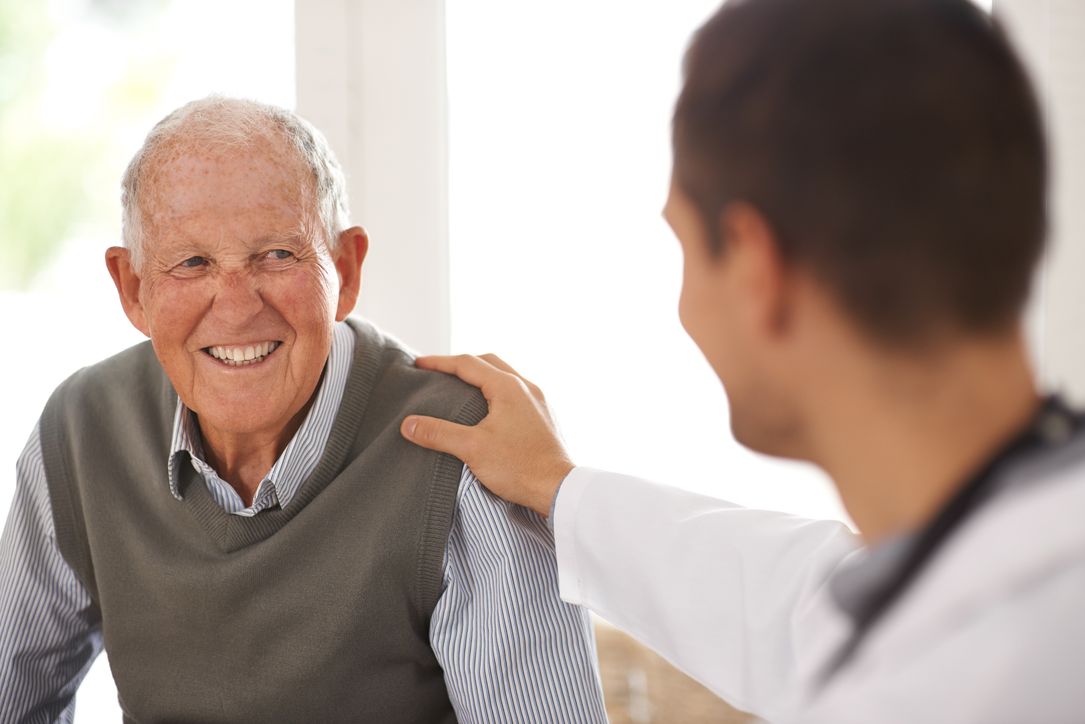 An elderly man smiling at a male physician in a white coat who has his hand on the elderly man's shoulder.