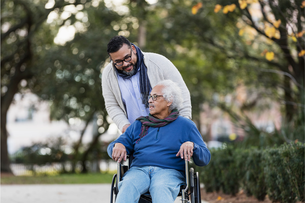 A son pushes his father in a wheelchair along a park path