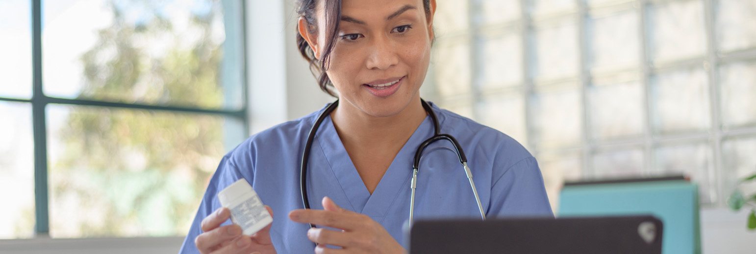 A nurse in scrubs points to a medicine bottle in front of a tablet.